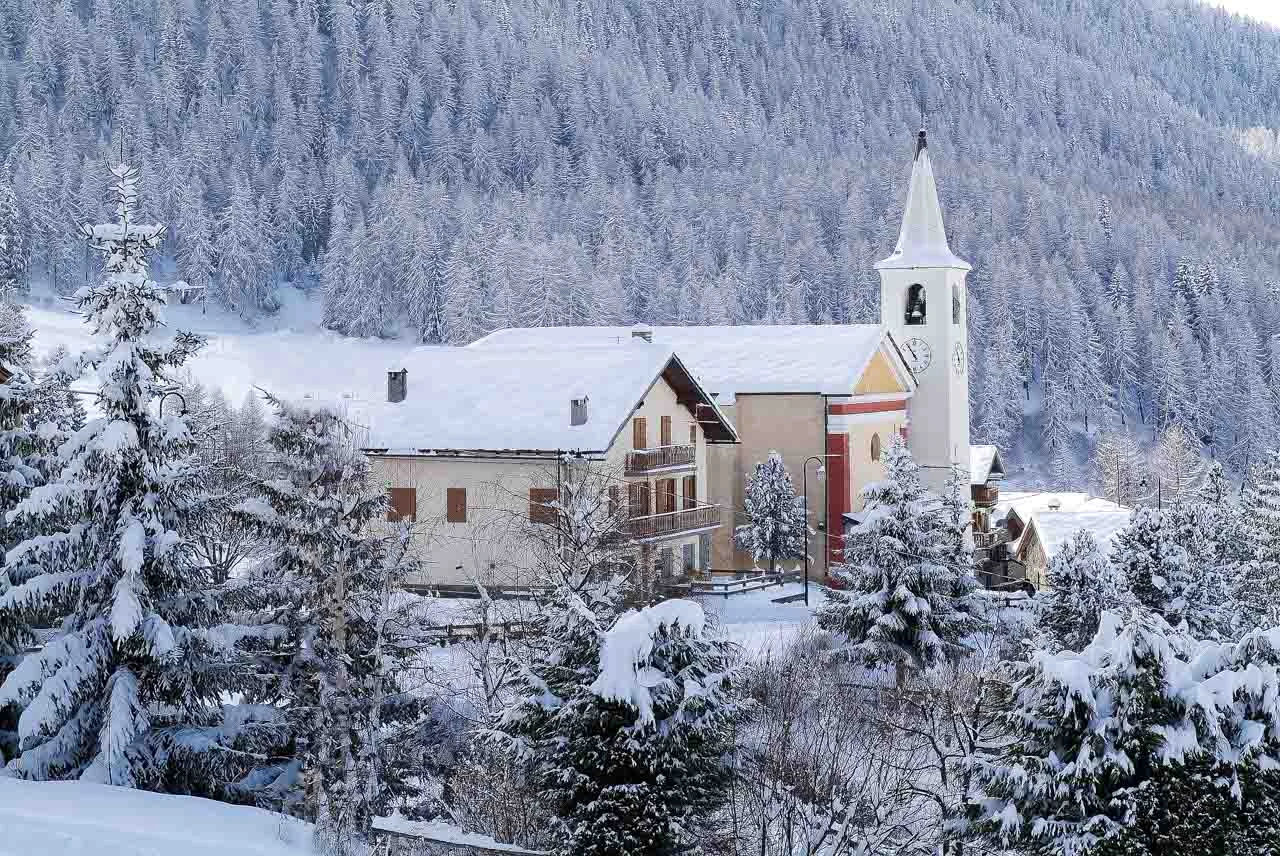 a church at the foot of a wooded mountain, covered in snow, in Aosta Valley