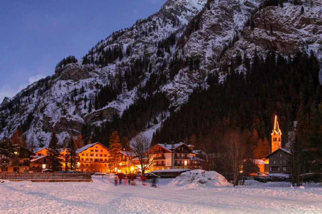 a church steeply and mountain village is lit up, at dusk, sitting under a snowy, rocky mountain side