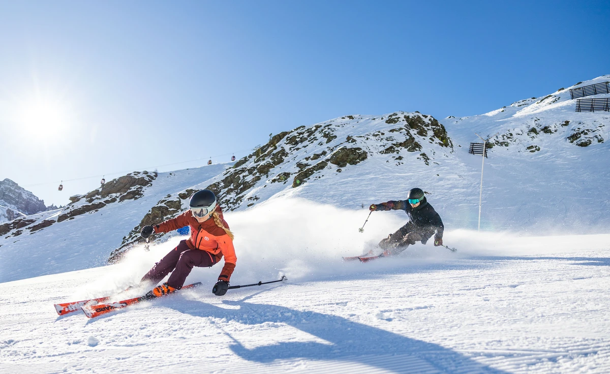 two skiers race down a groomed piste, clearly at speed, the second following closely in the first skier's tracks