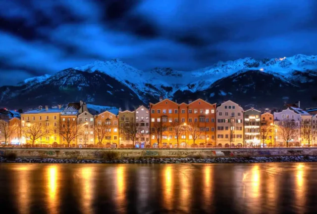 a colourful line of houses lit up, lights reflecting off the river in front, with snowy mountains as a backdrop