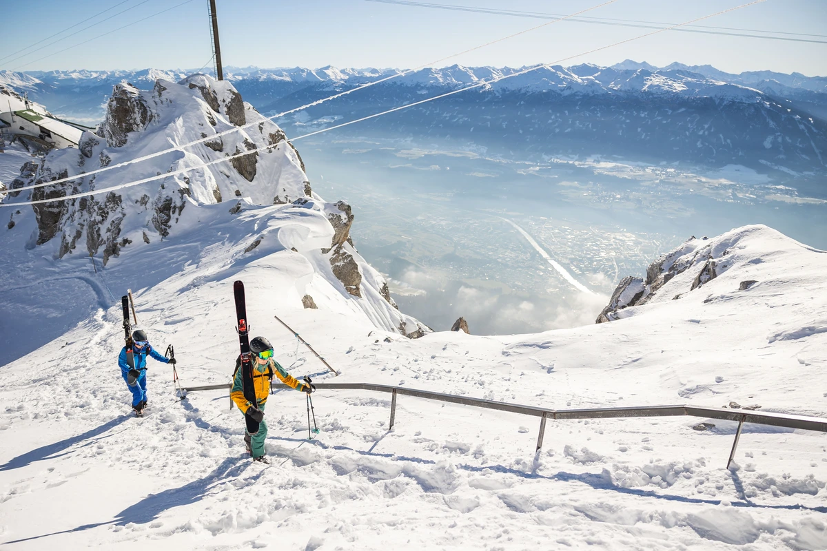 far below a river and city surrounded by snowy mountains, with two skiers hiking uphill, skis on back, in the foreground