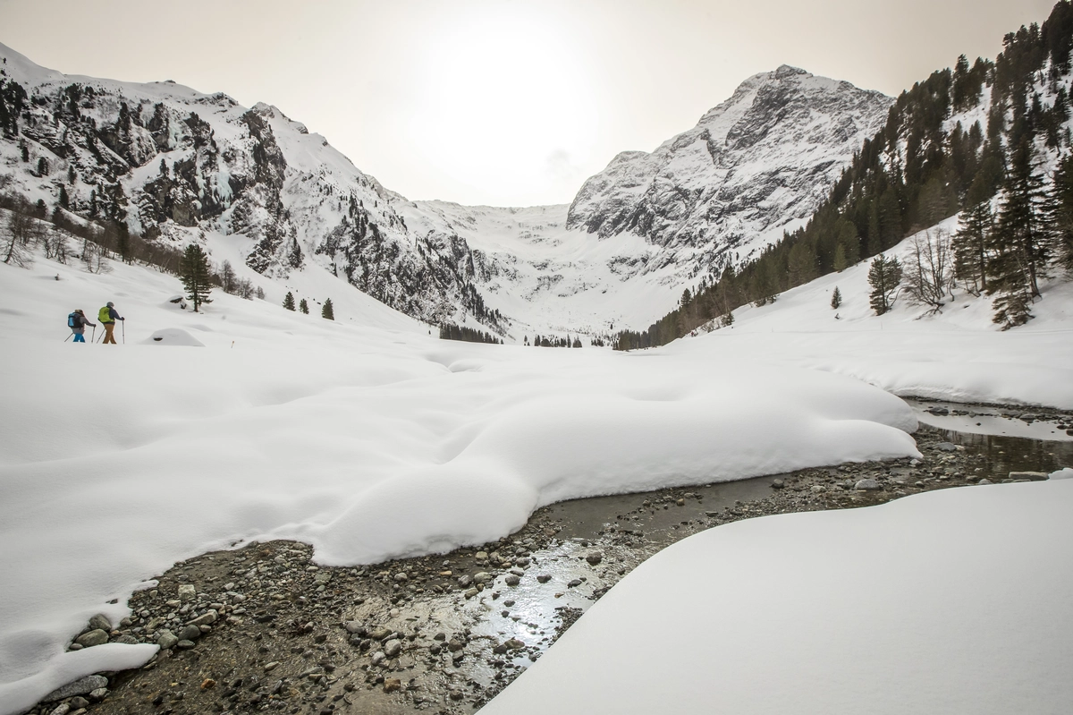 two snowshoers hike through a valley covered in snow besid a river